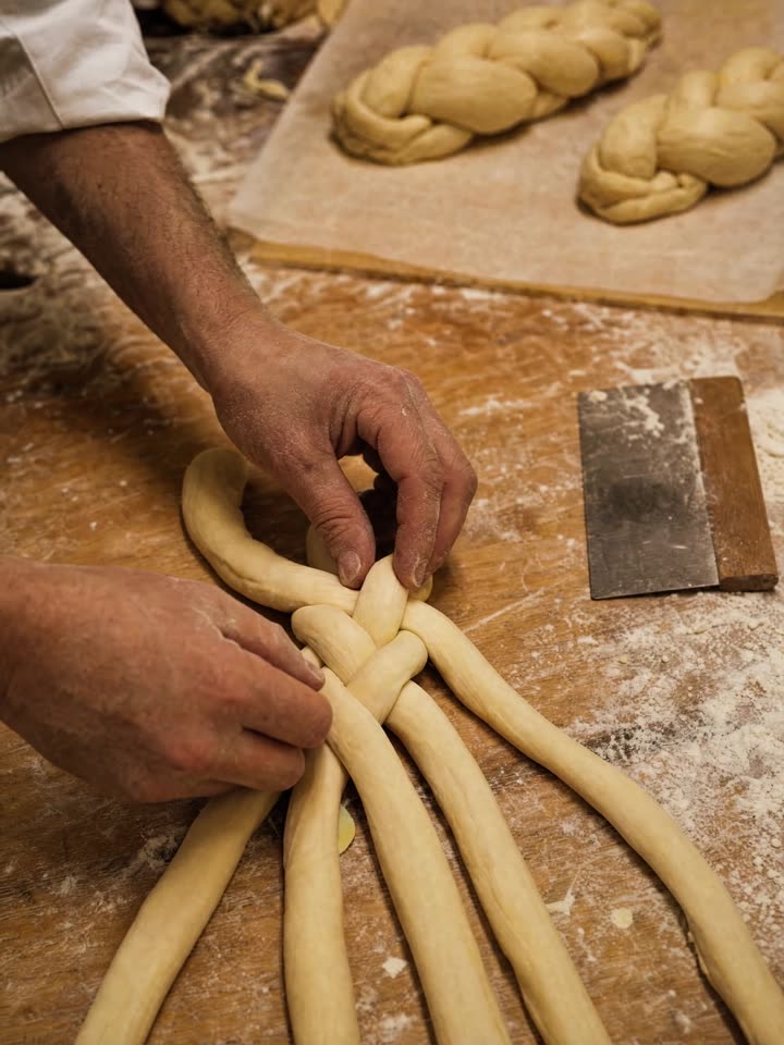 Hands braiding strands of challah dough on a floured bench